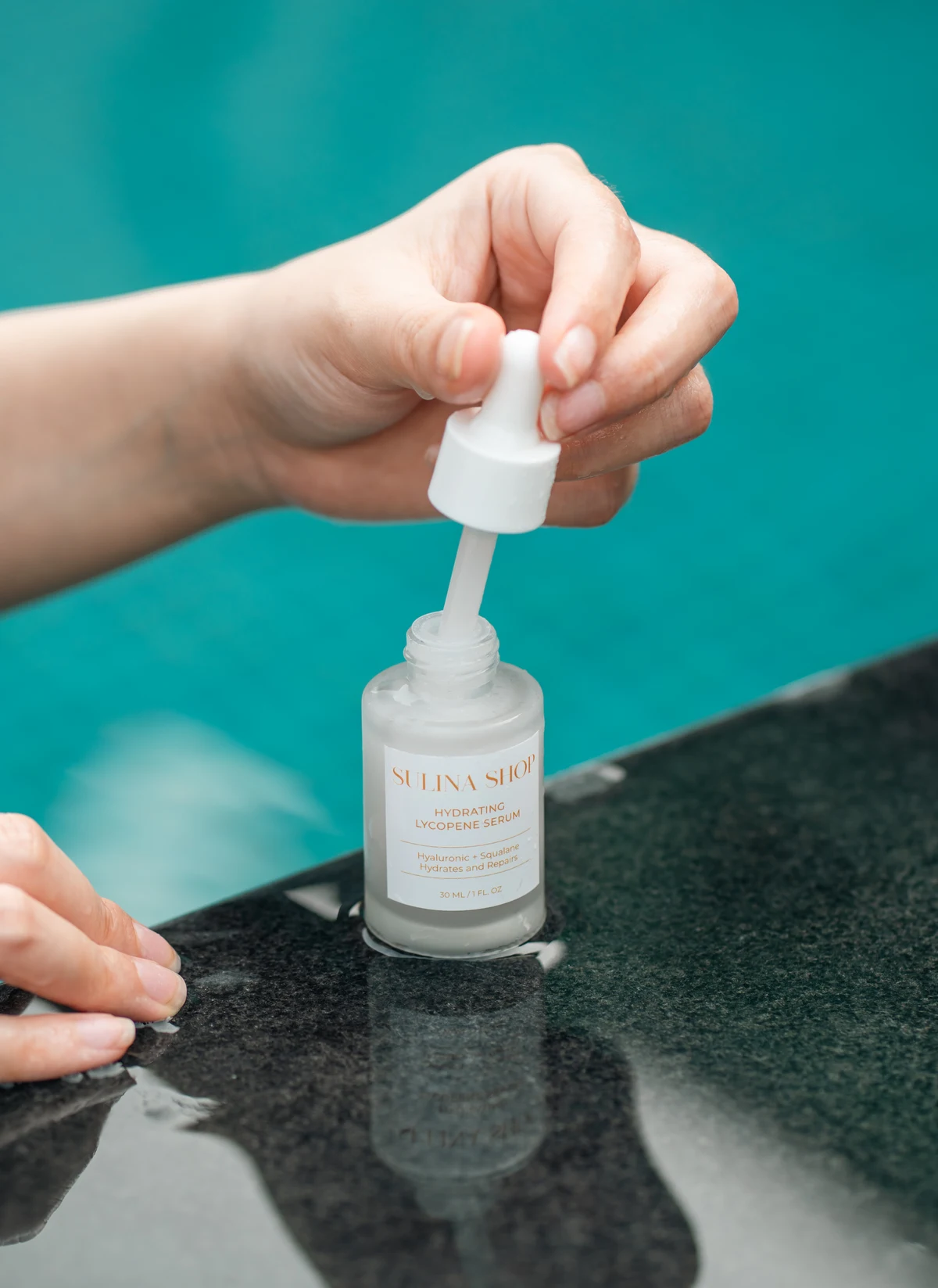 Hands opening a Sulina Shop serum bottle beside a turquoise pool